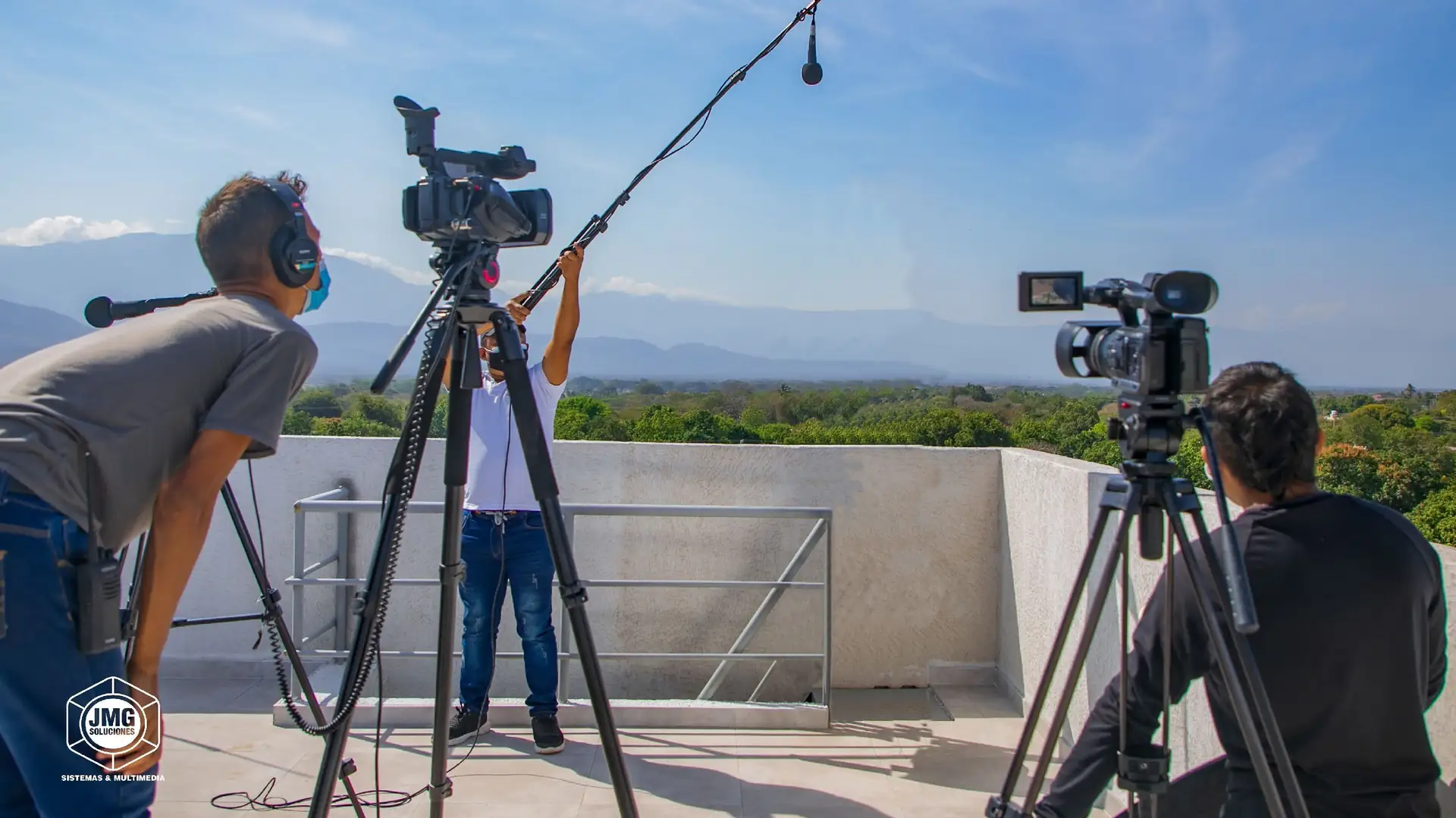 Equipo de JMG graba en una azotea con dos cámaras de video sobre trípodes y un micrófono de caña, con montañas al fondo.
