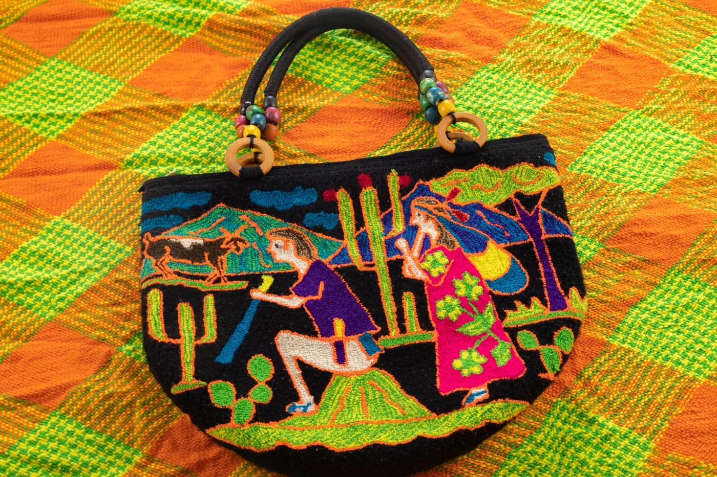 Bolso negro bordado con figuras de colores sobre una tela a cuadros verde y naranja.