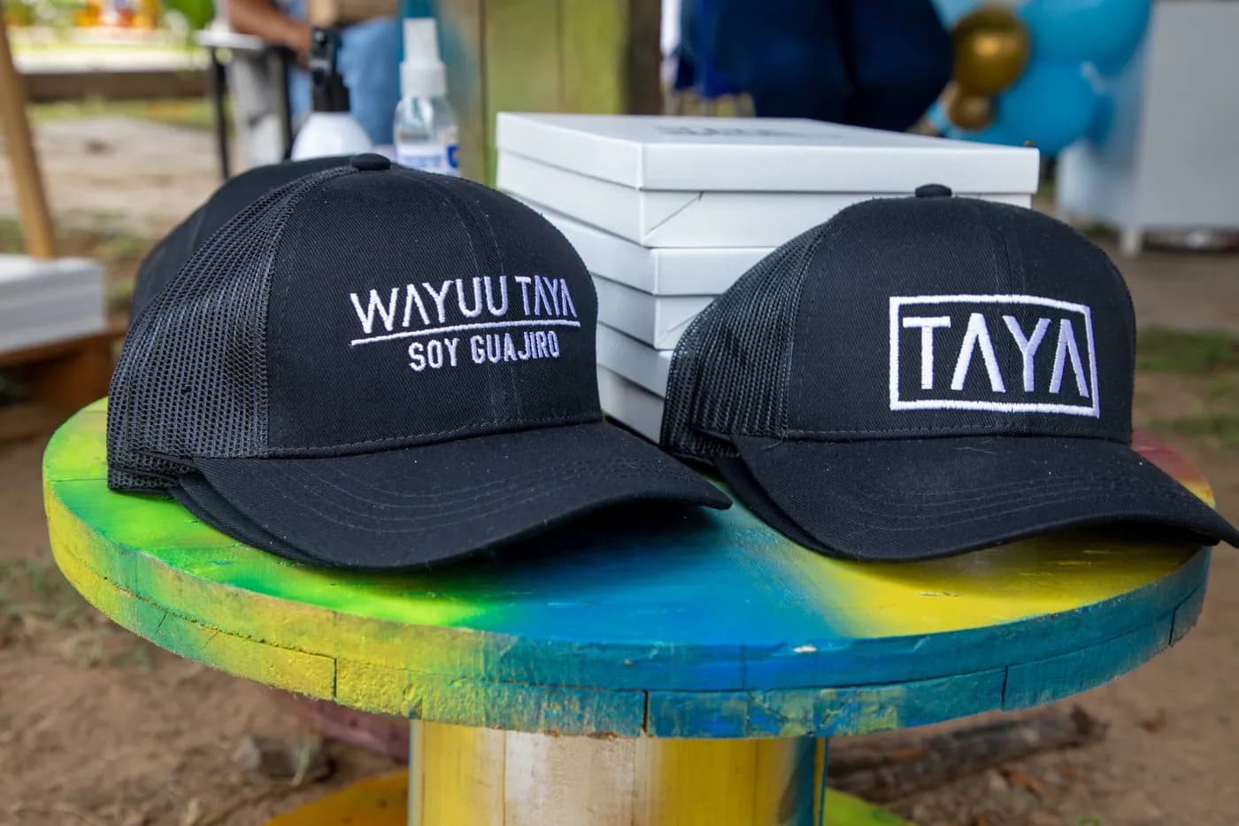 Dos gorras negras con marca bordada sobre una mesa redonda pintada de colores.