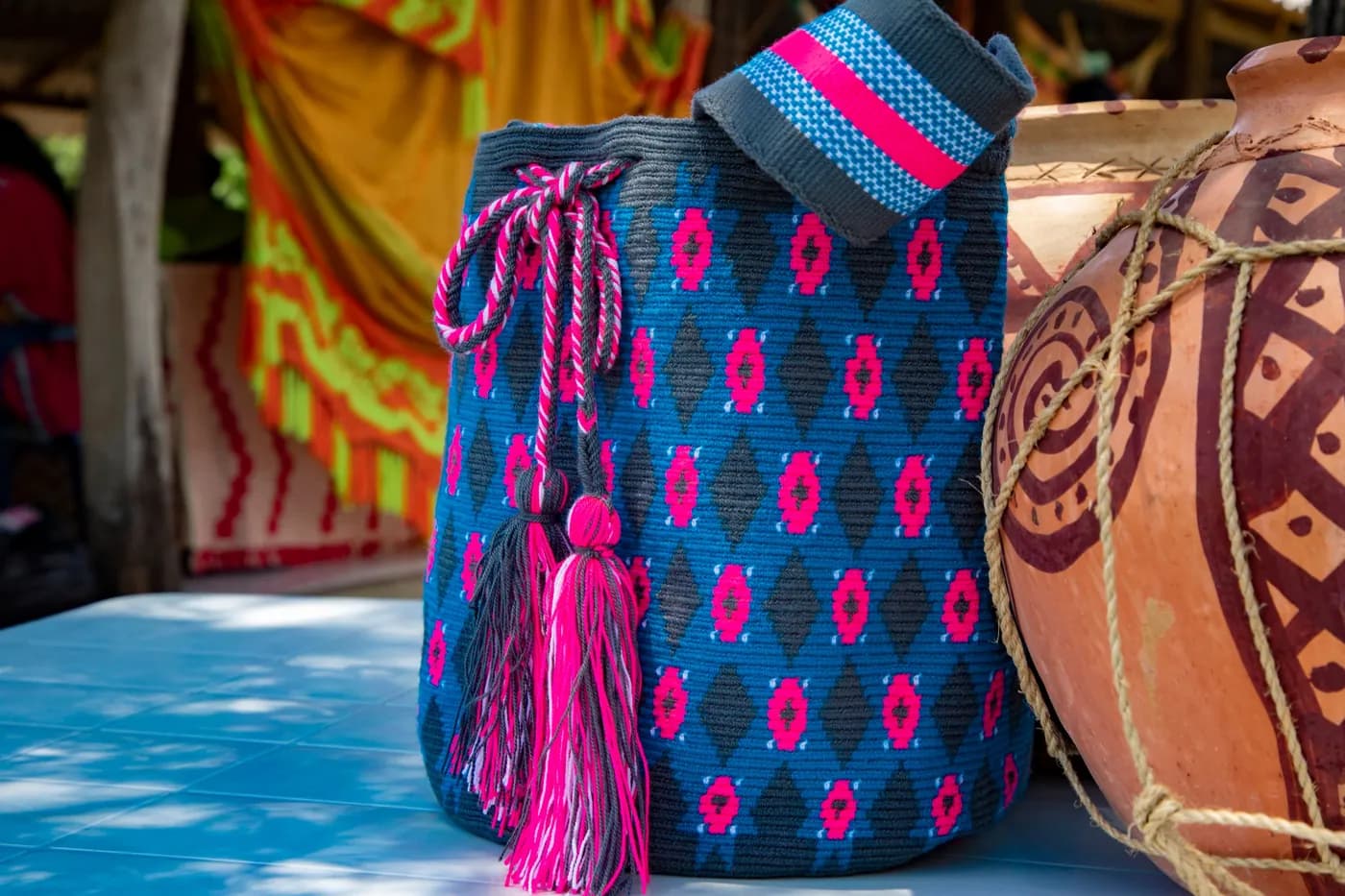 Mochila tejida azul y fucsia junto a vasijas de barro decoradas.