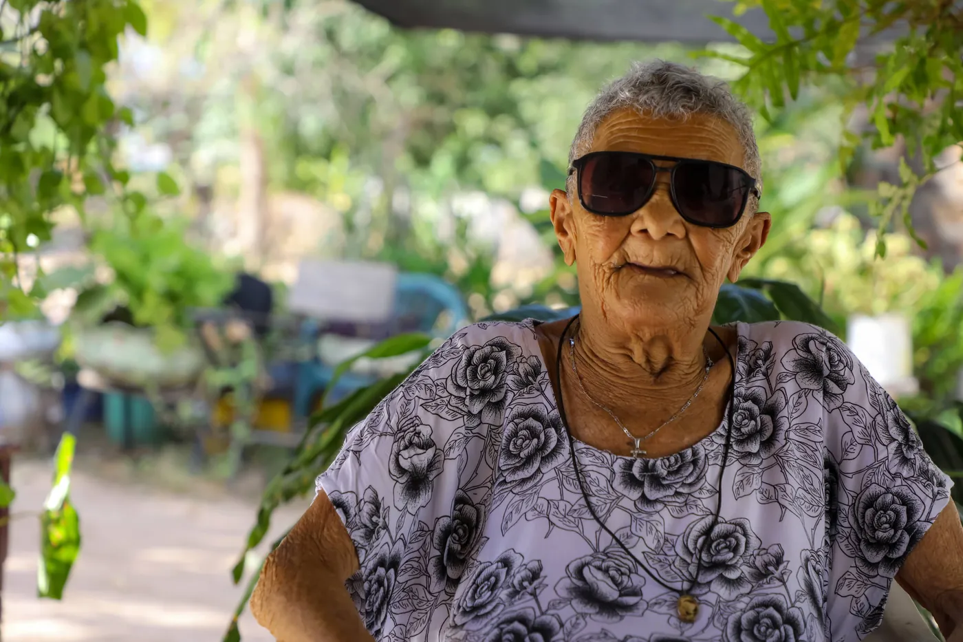 Retrato de una mujer mayor con gafas oscuras y blusa de flores en un patio con árboles.