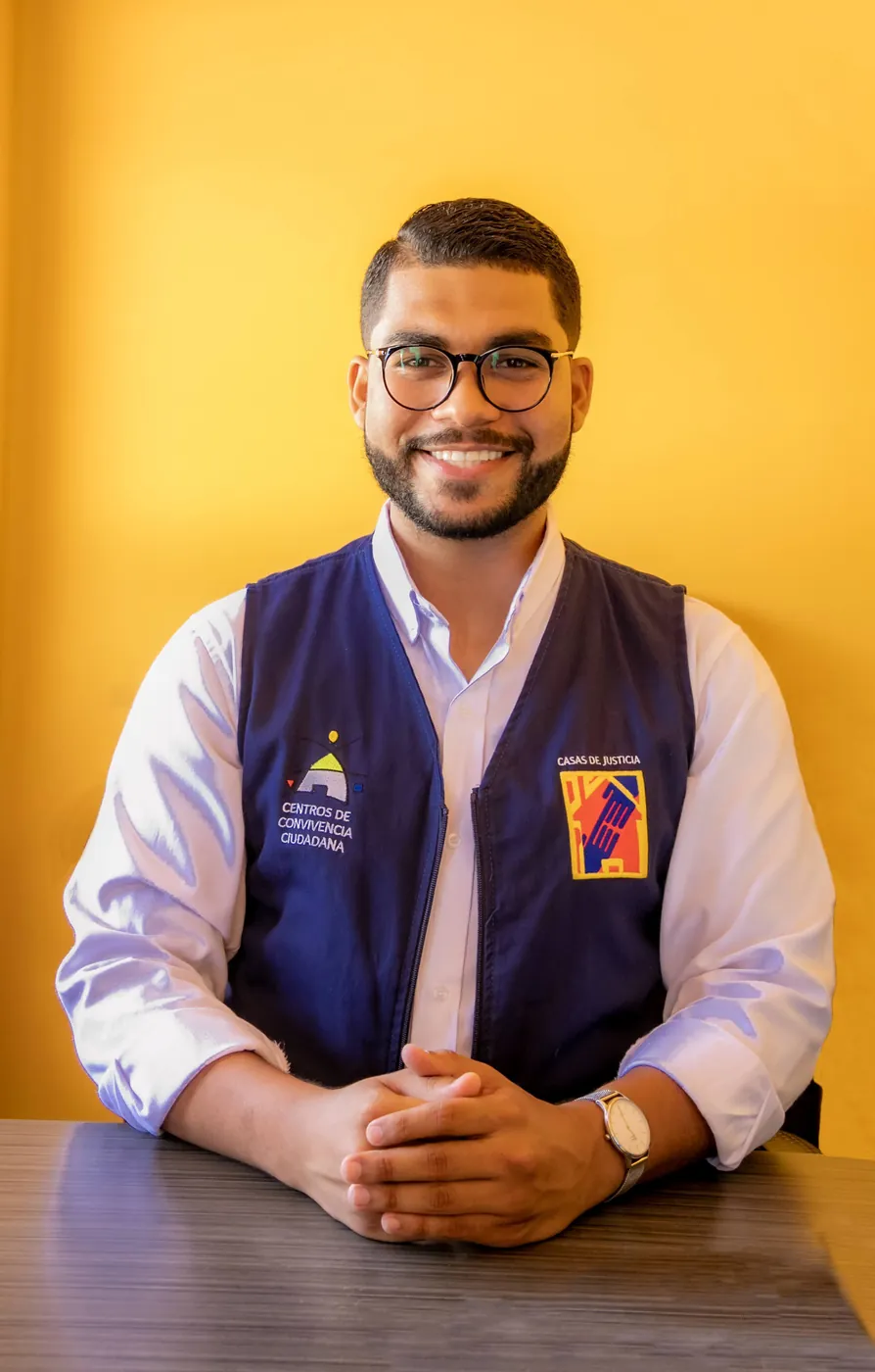 Retrato institucional de un hombre con chaleco azul sentado ante una mesa, sobre un fondo amarillo.