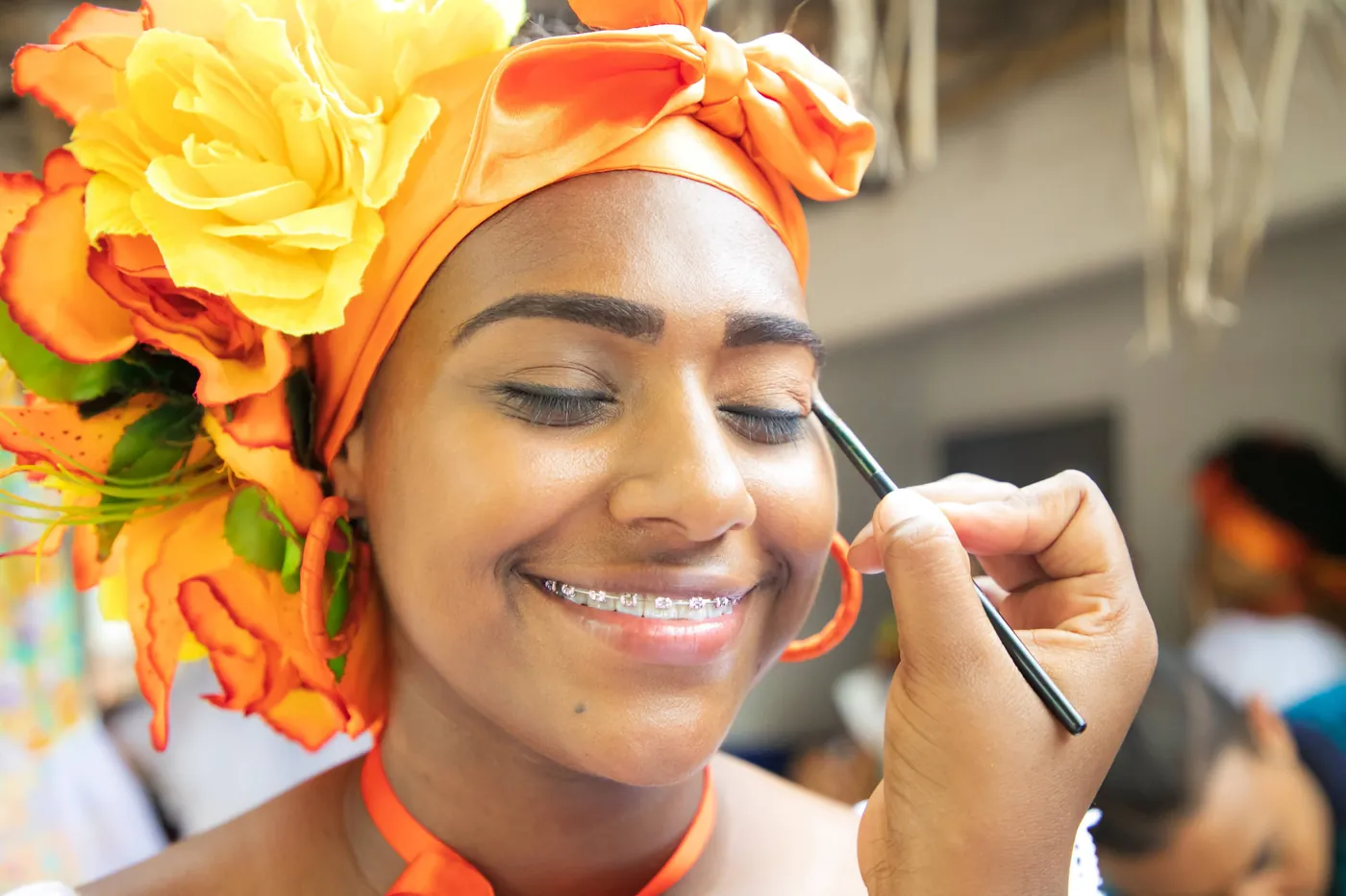 Maquilladora aplica color a una mujer con un tocado de flores naranjas y amarillas.