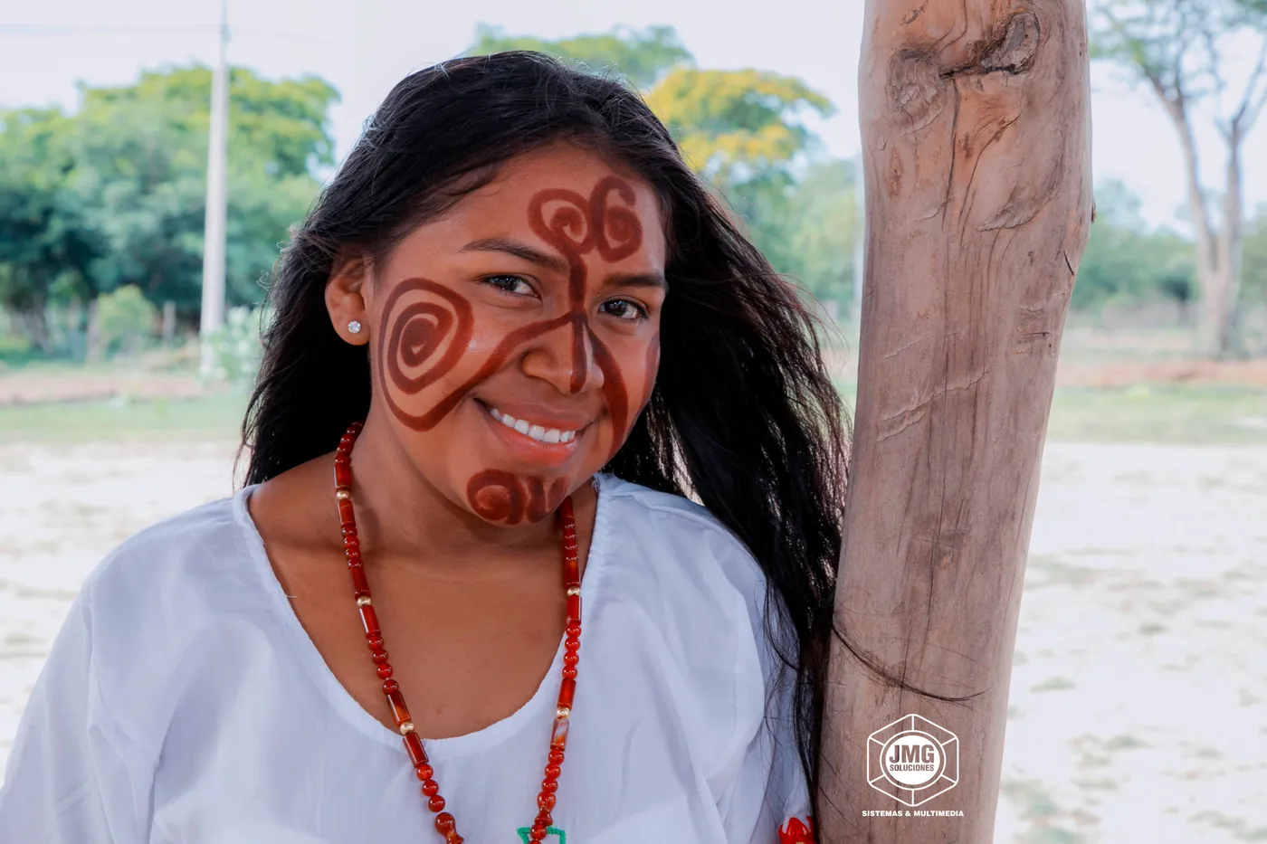 Retrato de una mujer sonriente con pintura facial tradicional en tonos rojizos y collar de cuentas.