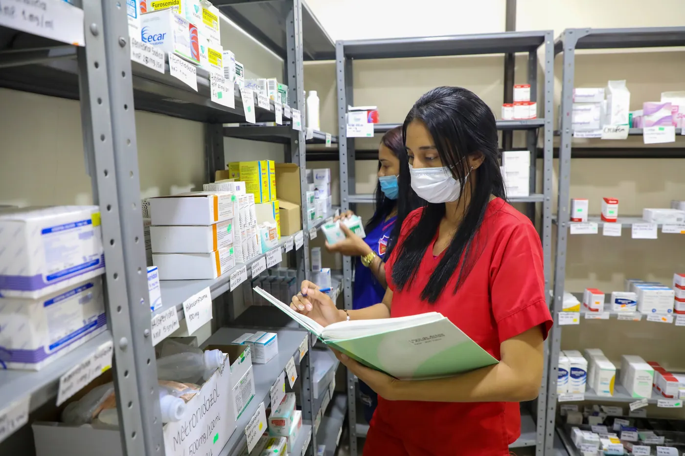 Auxiliar de farmacia con tapabocas revisa un registro entre estanterías de medicamentos.