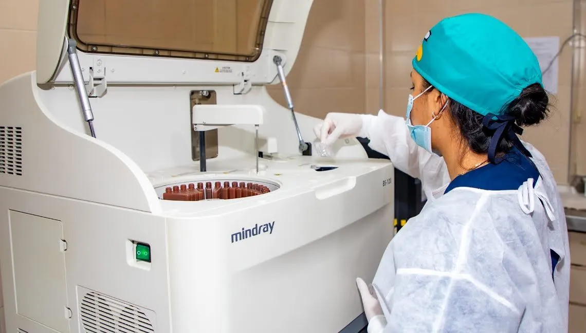 Bacterióloga con gorro y bata carga muestras en un equipo de análisis de laboratorio.