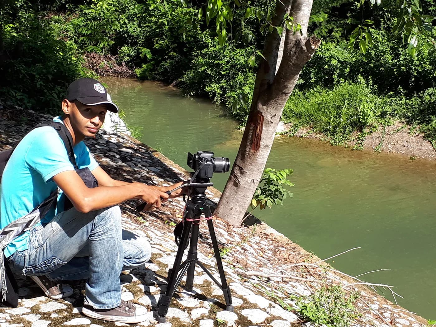 Camarógrafo de JMG ajusta una cámara sobre trípode a la orilla de un río.
