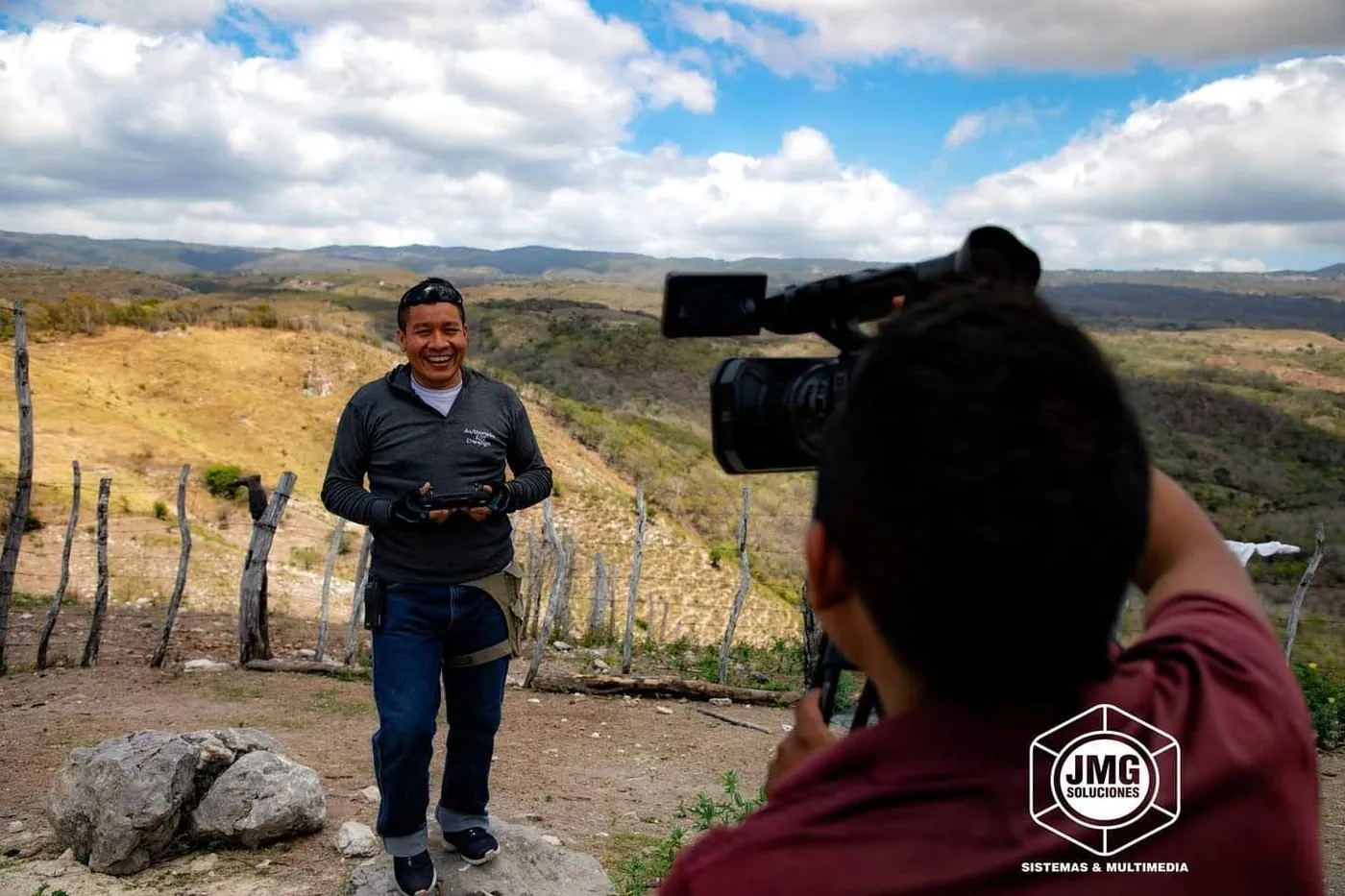 Camarógrafo de JMG graba a un hombre que habla en un campo seco con colinas al fondo.