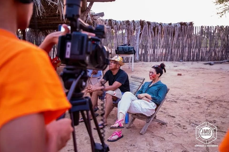 Camarógrafo de JMG graba una entrevista a dos personas sentadas en una ranchería, frente a un corral de madera.