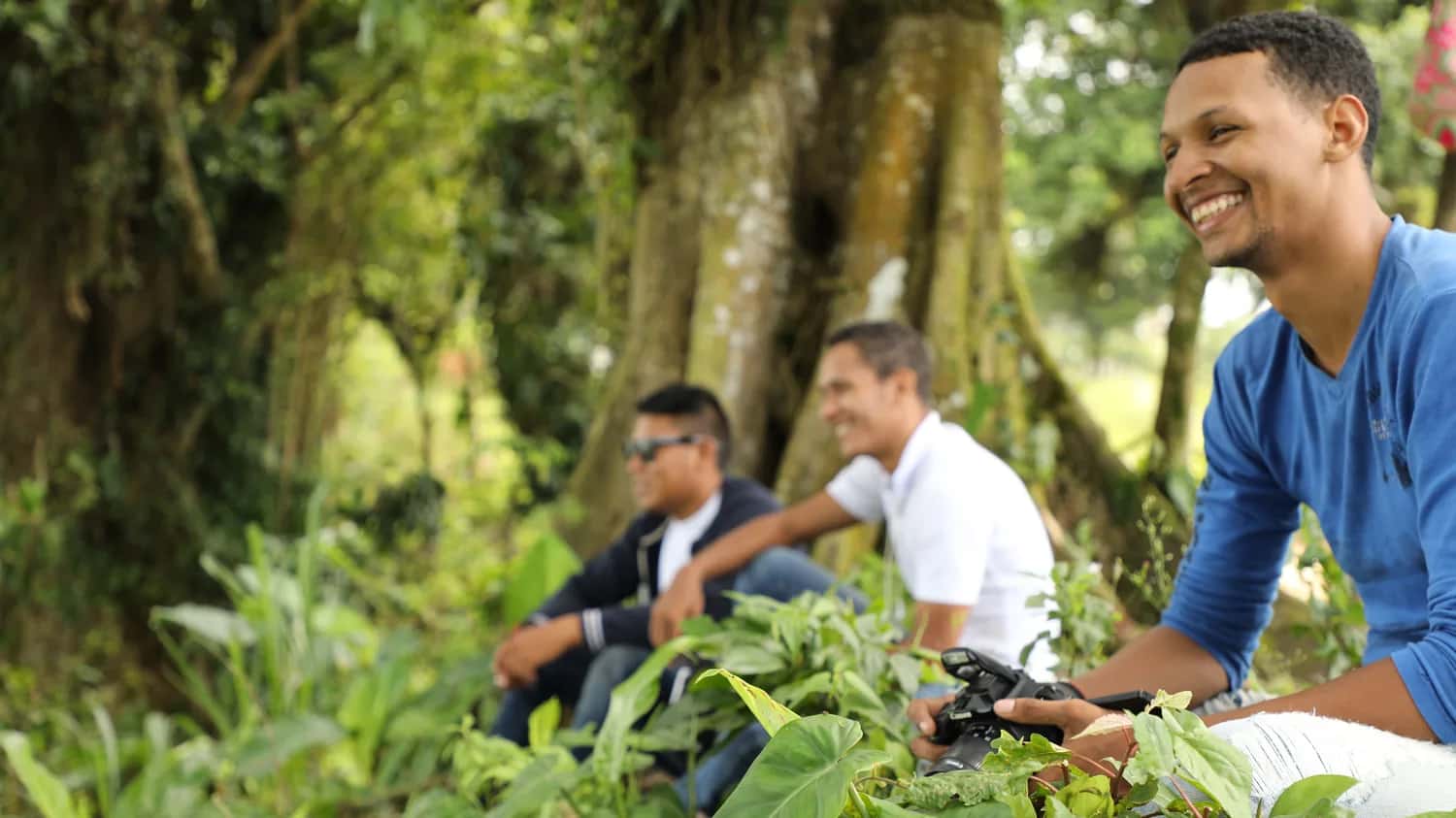 Tres jóvenes conversan y ríen sentados bajo los árboles; uno sostiene una cámara fotográfica.