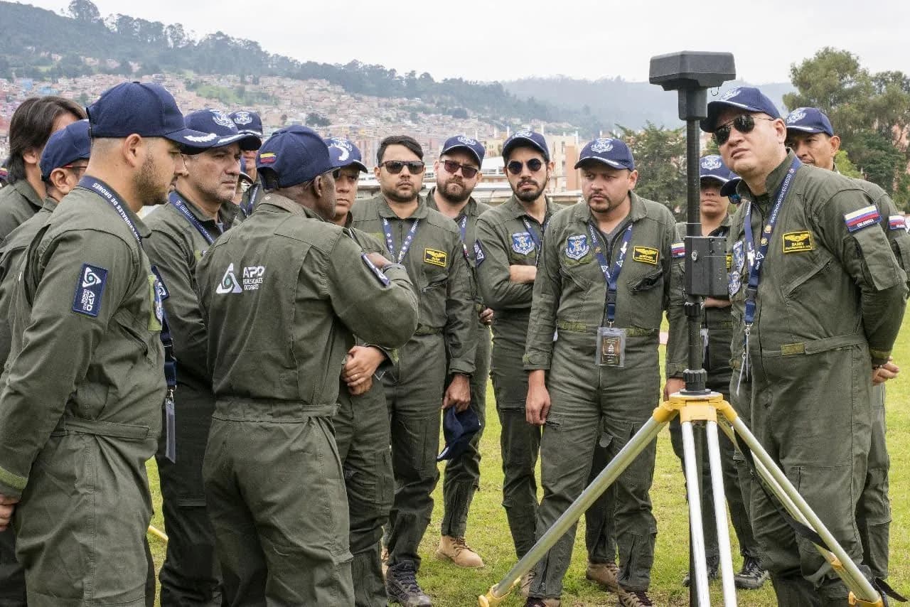 Grupo de pilotos con overol de vuelo escucha la explicación de una estación base GNSS montada sobre un trípode.