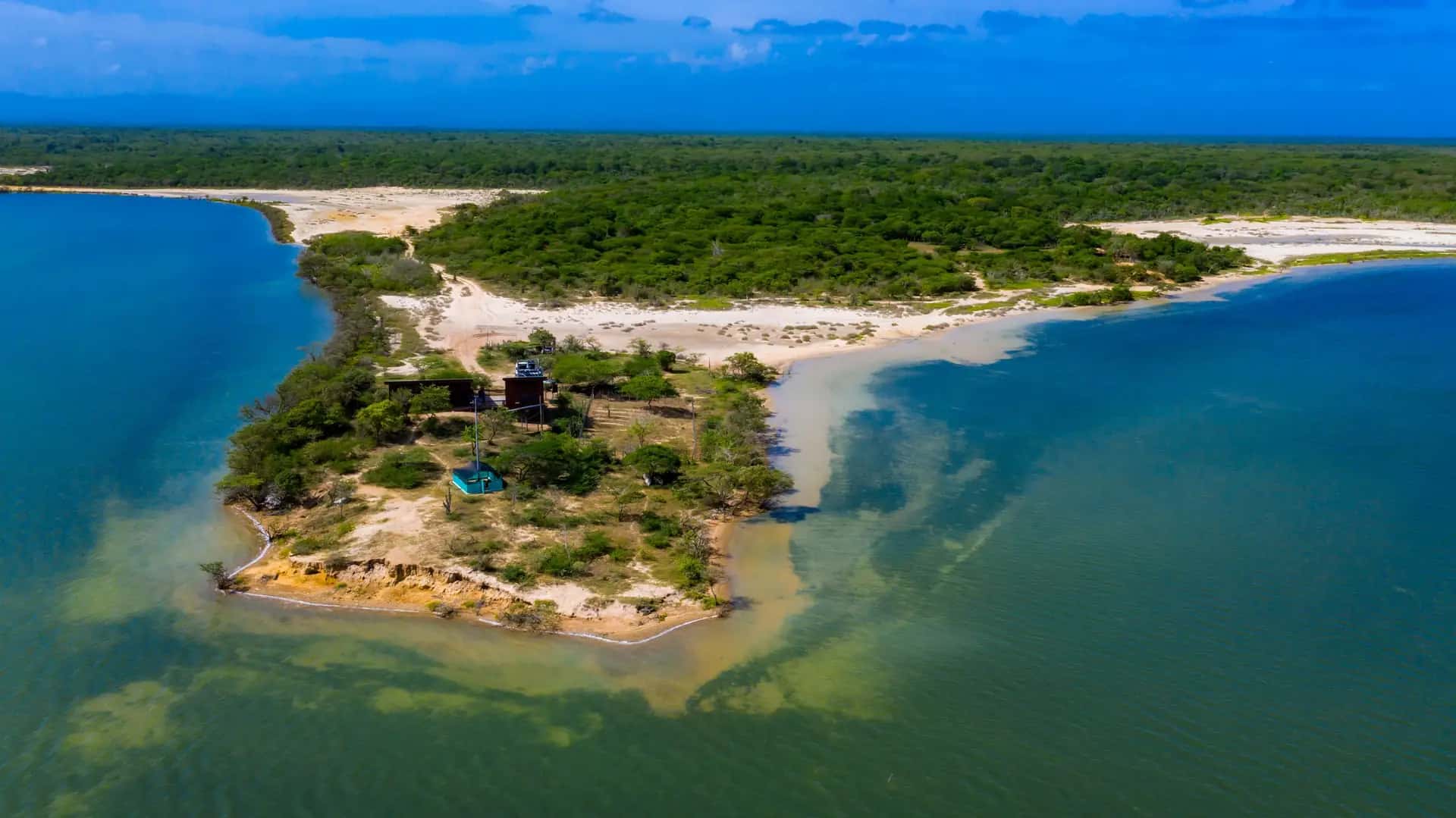 Vista aérea con dron de una isla de arena y vegetación rodeada de aguas de distintos tonos de azul, junto a la costa.
