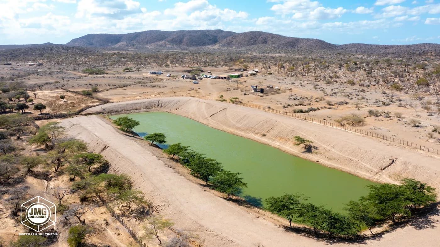 Vista aérea con dron de un reservorio de agua rodeado de árboles en una zona semidesértica, con montañas al fondo.