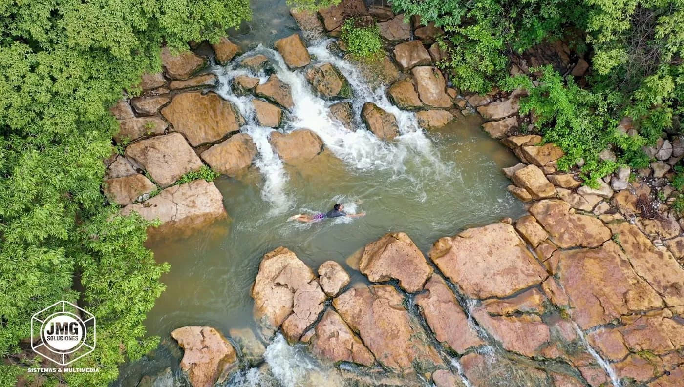 Vista cenital con dron de una cascada que corre entre grandes rocas rojizas y vegetación.