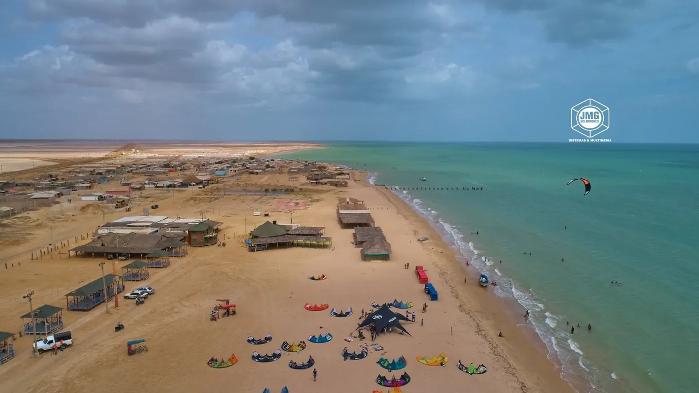 Vista aérea con dron de una playa con kioscos y cometas de kitesurf sobre la arena, junto a un mar verde turquesa.