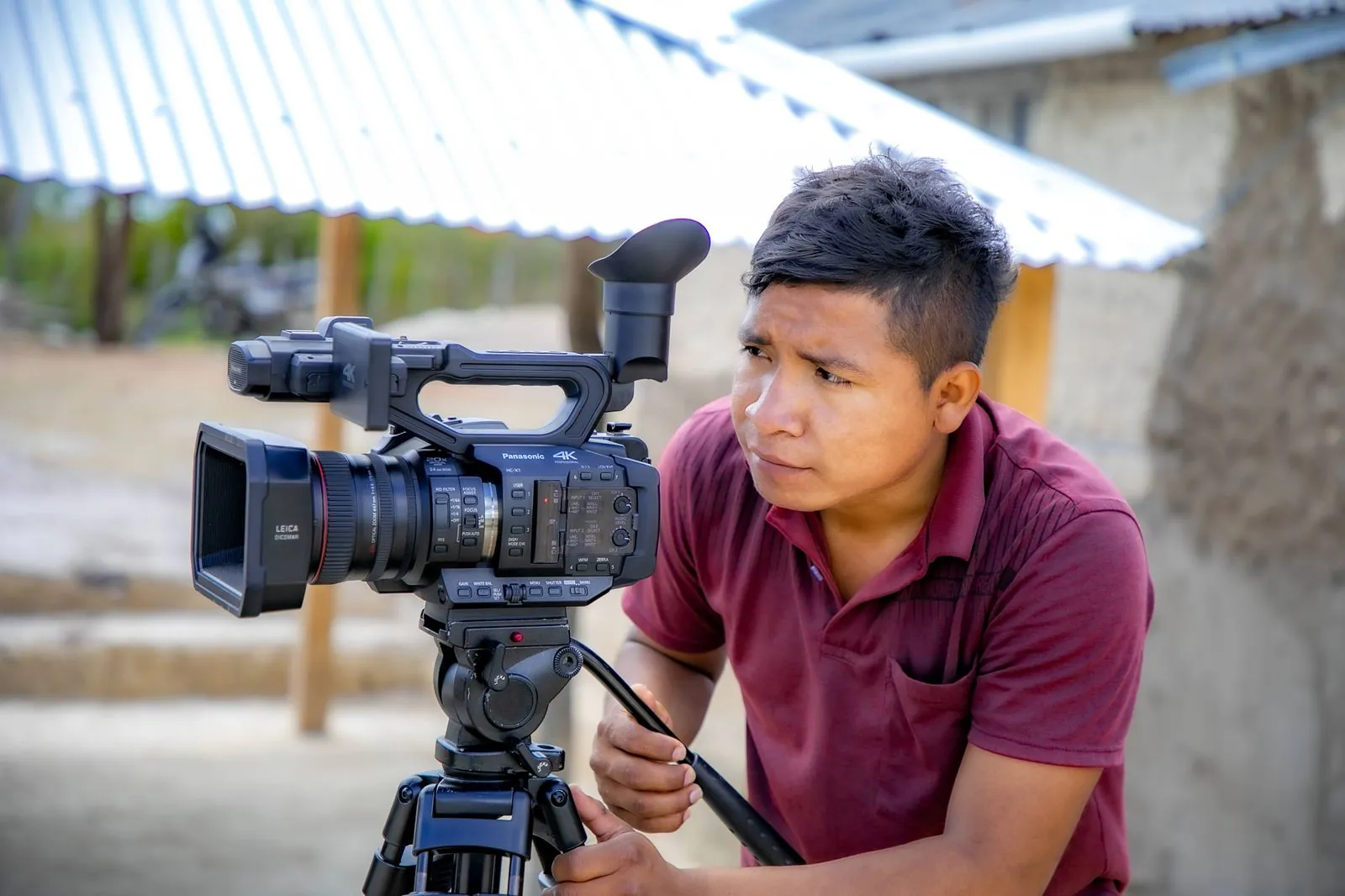 Camarógrafo de JMG encuadra con una cámara de video sobre trípode frente a una casa de bahareque con techo de zinc.