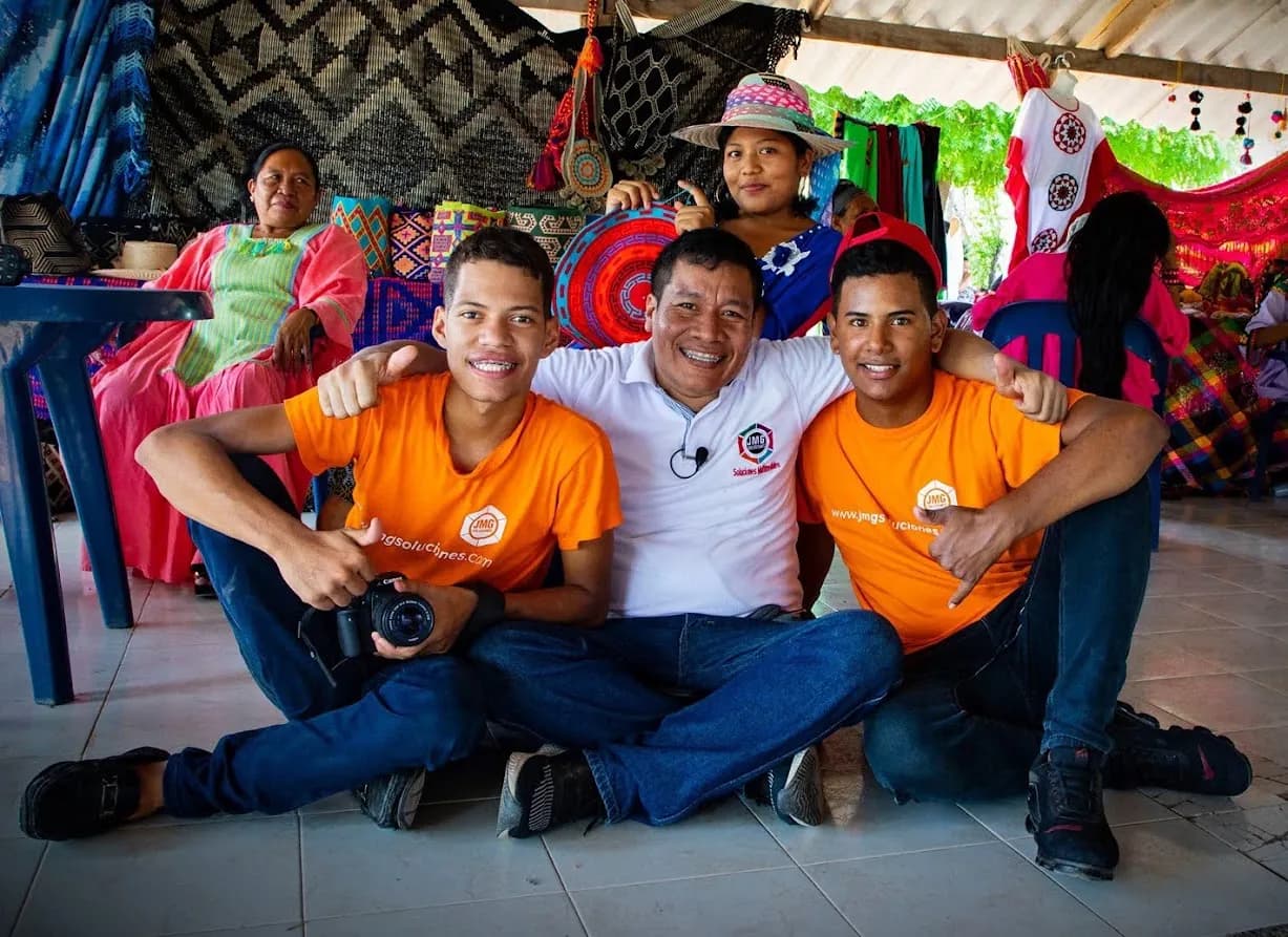 Integrantes de JMG con camisetas corporativas y cámara, sentados junto a artesanas wayuu y sus tejidos.