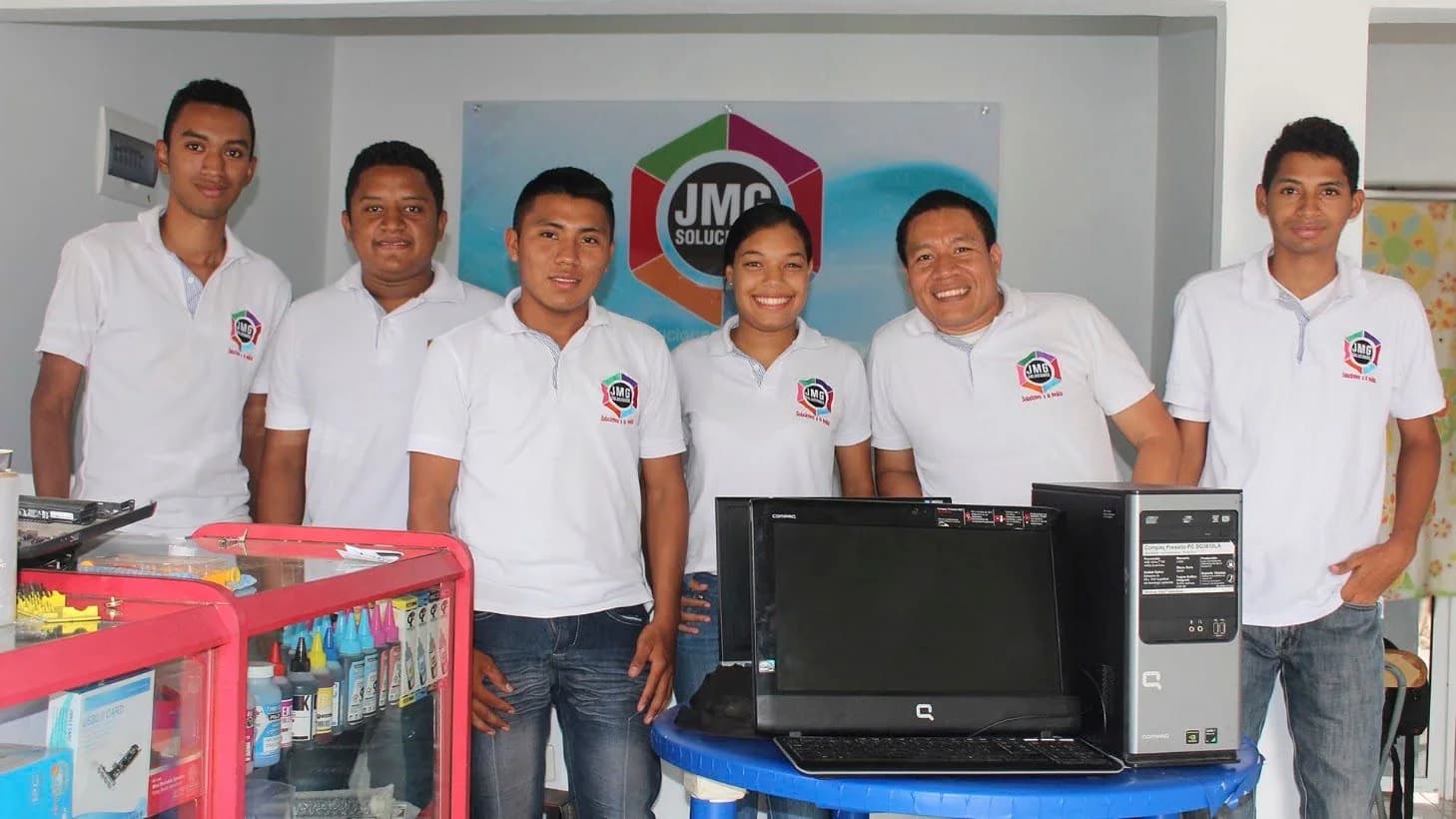 Equipo de JMG con camisetas blancas en el local, junto a computadores y una vitrina de insumos.