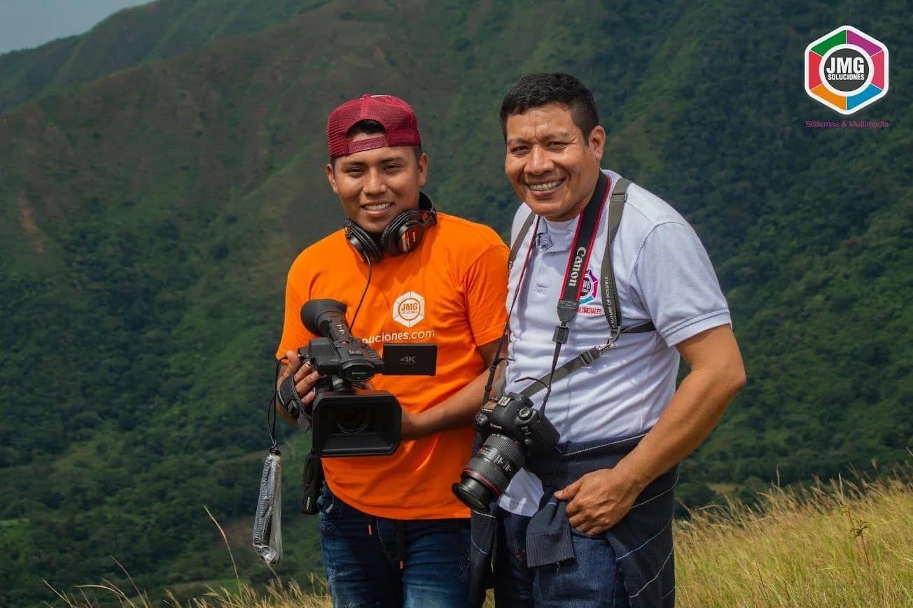 Dos integrantes de JMG sonríen en la montaña con una cámara de video y una cámara fotográfica.