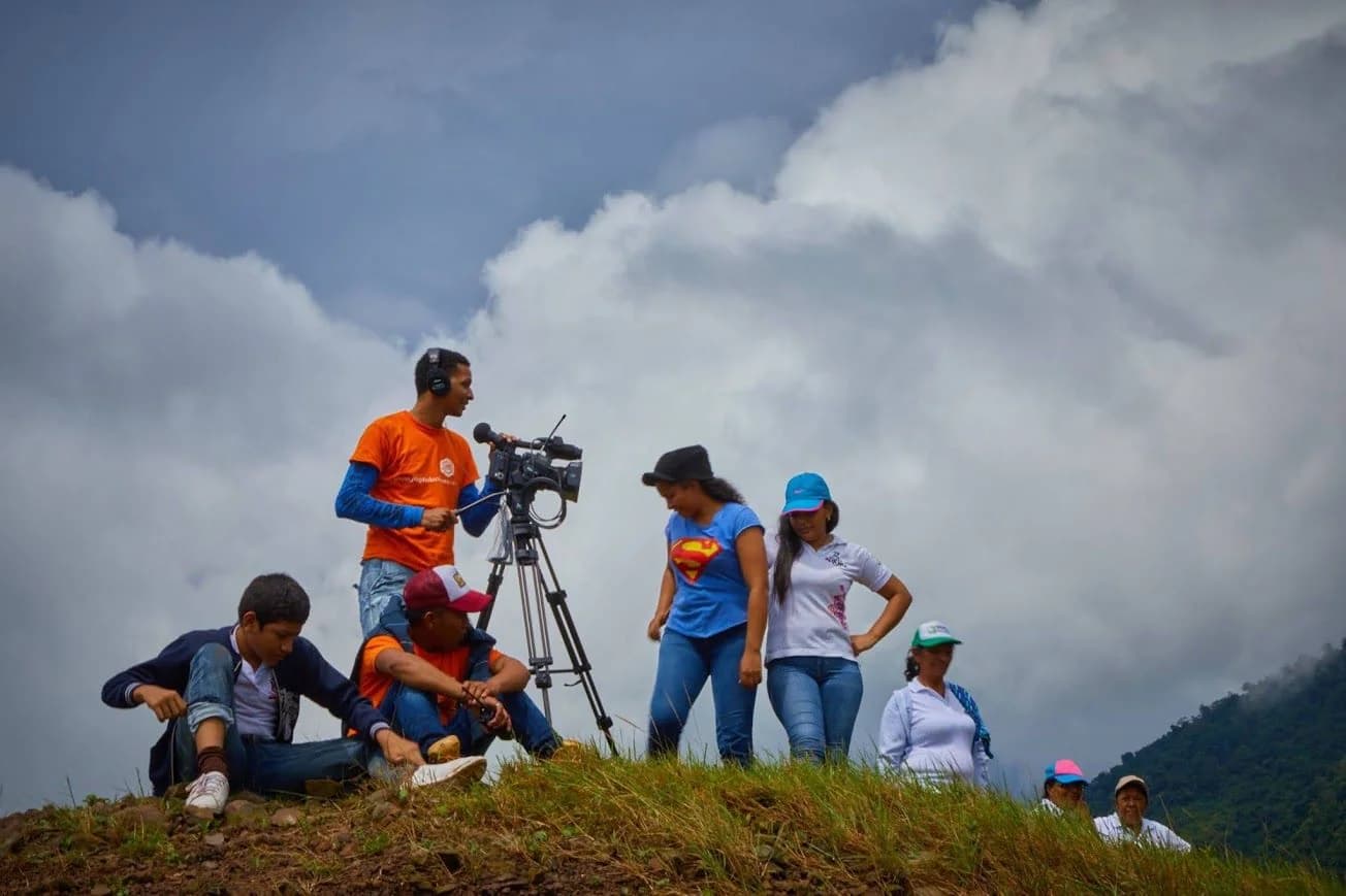 Camarógrafo de JMG graba con cámara sobre trípode en lo alto de una colina, rodeado de participantes, bajo un cielo de nubes.
