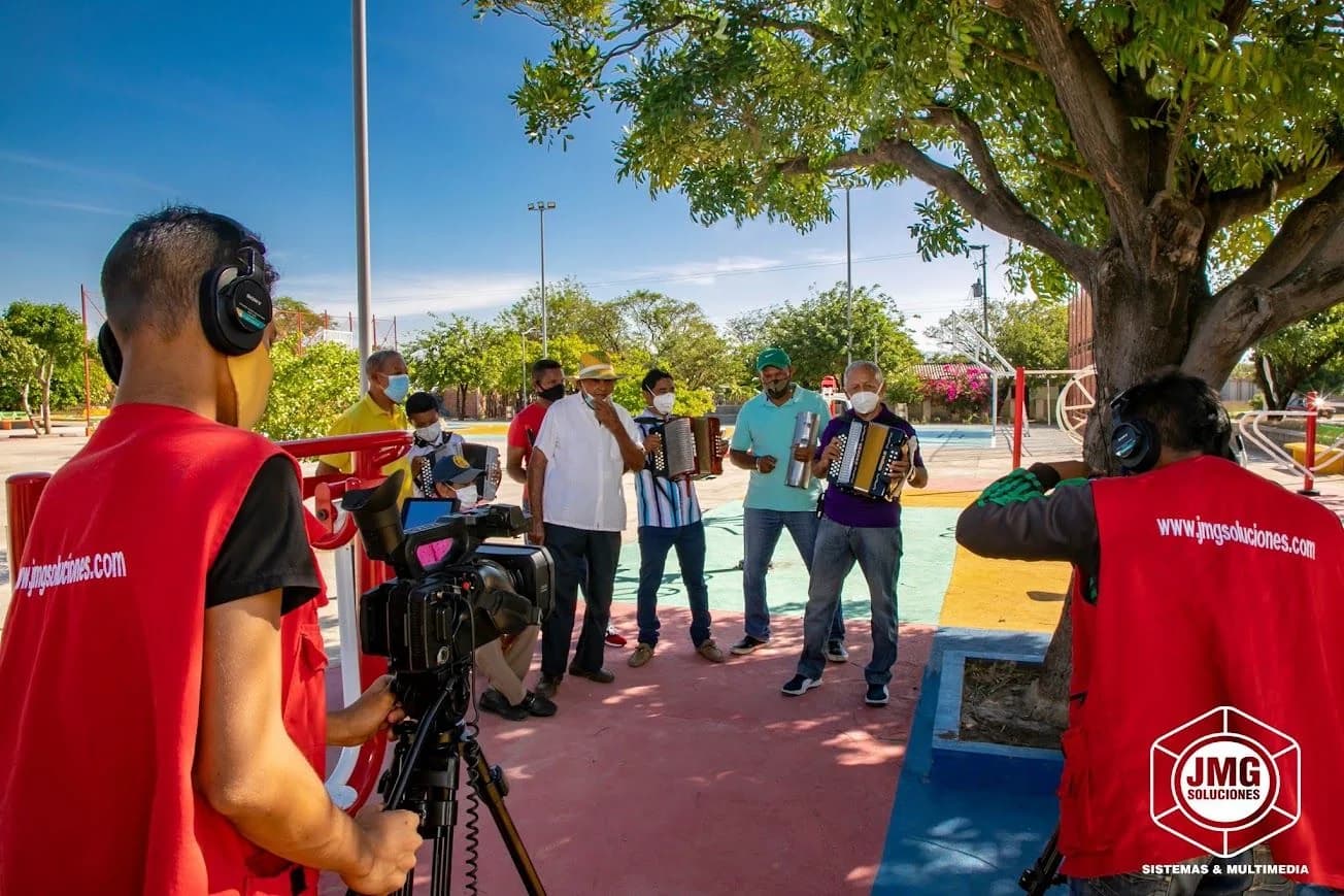 Dos operadores de JMG con chalecos rojos graban con cámara y audífonos a un grupo de músicos de acordeón en un parque.