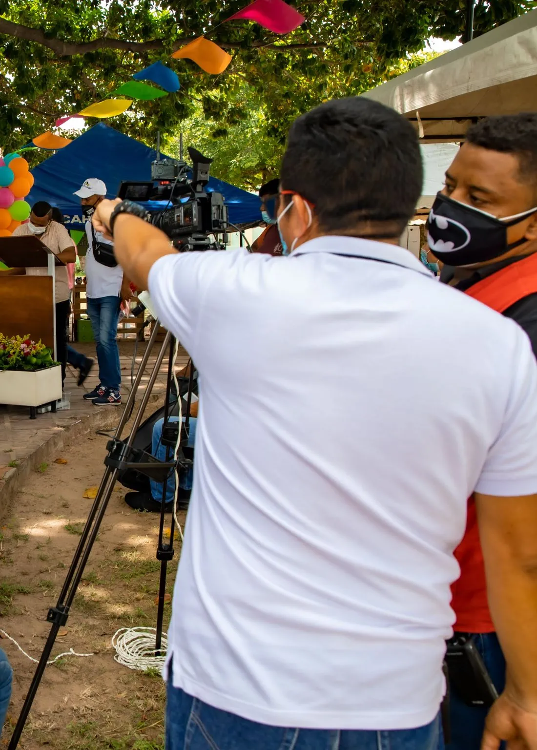Camarógrafo de JMG con chaleco corporativo graba con cámara sobre trípode durante una feria de emprendedores.