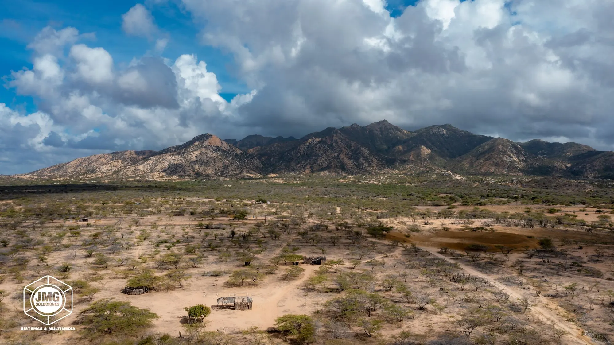 Vista aérea del monte seco de La Guajira con una enramada tradicional y la serranía al fondo bajo nubes de tormenta.