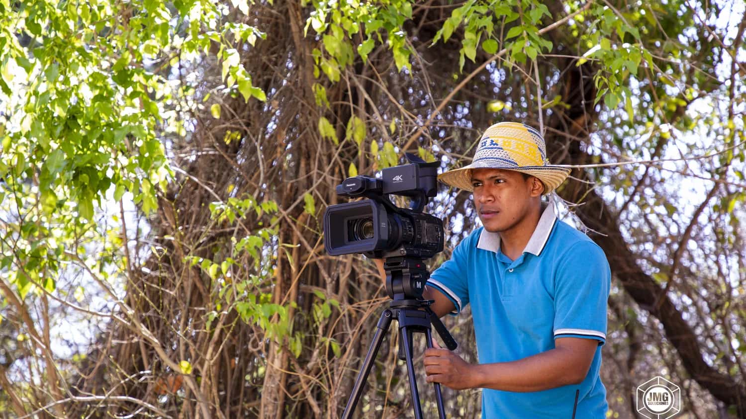 Camarógrafo de JMG con sombrero vueltiao opera una cámara 4K sobre trípode bajo los árboles de La Guajira.