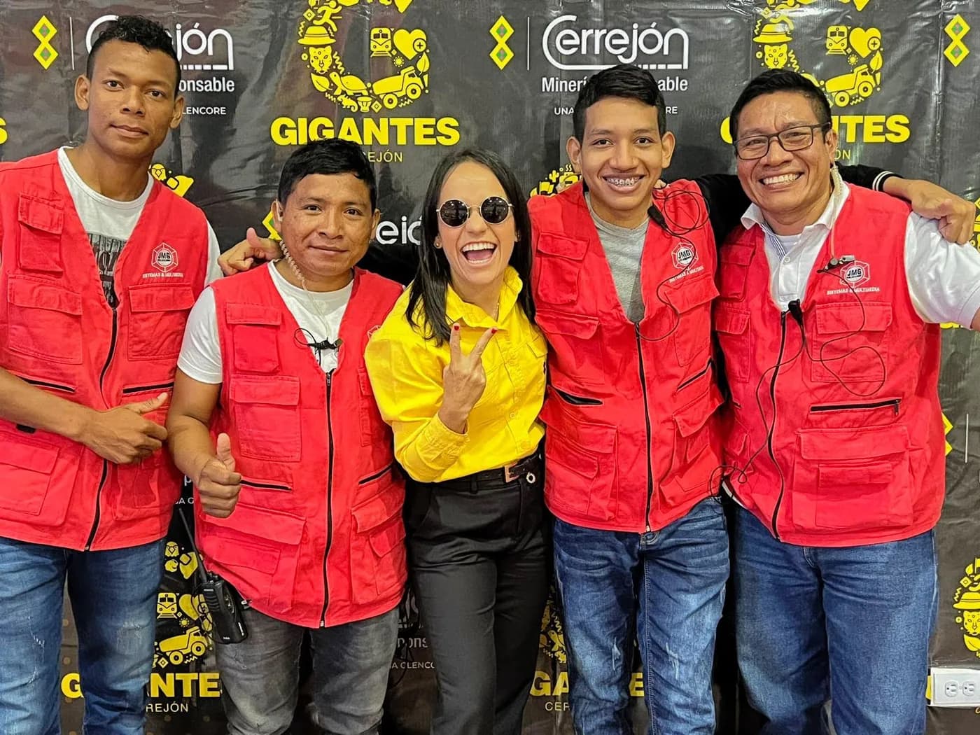 Integrantes de JMG con chalecos rojos posan con la presentadora del programa «Gigantes» frente al fondo de prensa de Cerrejón.