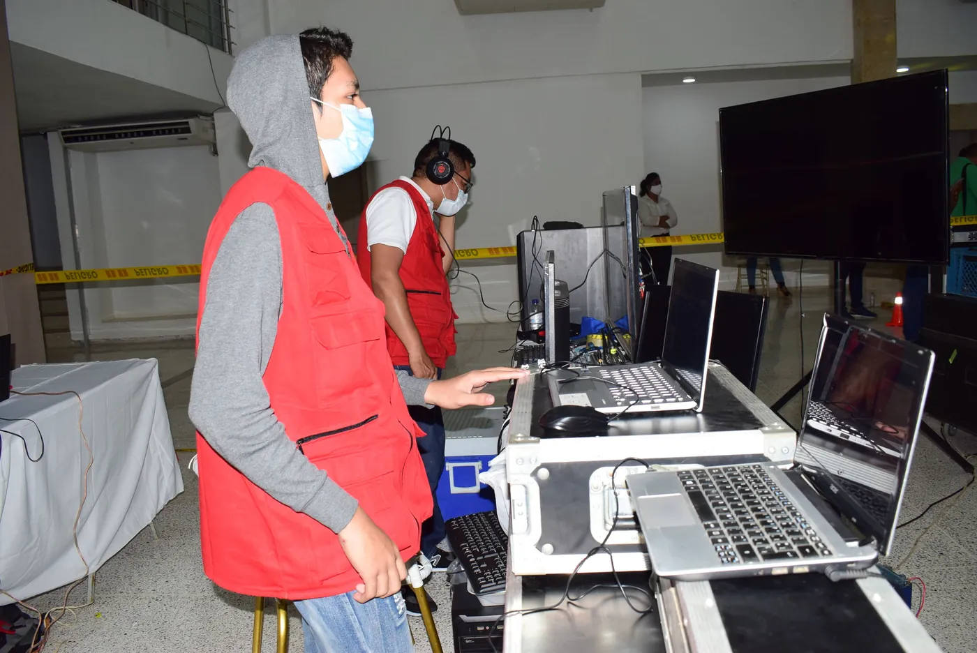 Dos operadores de JMG con chalecos rojos y audífonos trabajan en la mesa de control, con portátiles y una pantalla grande.