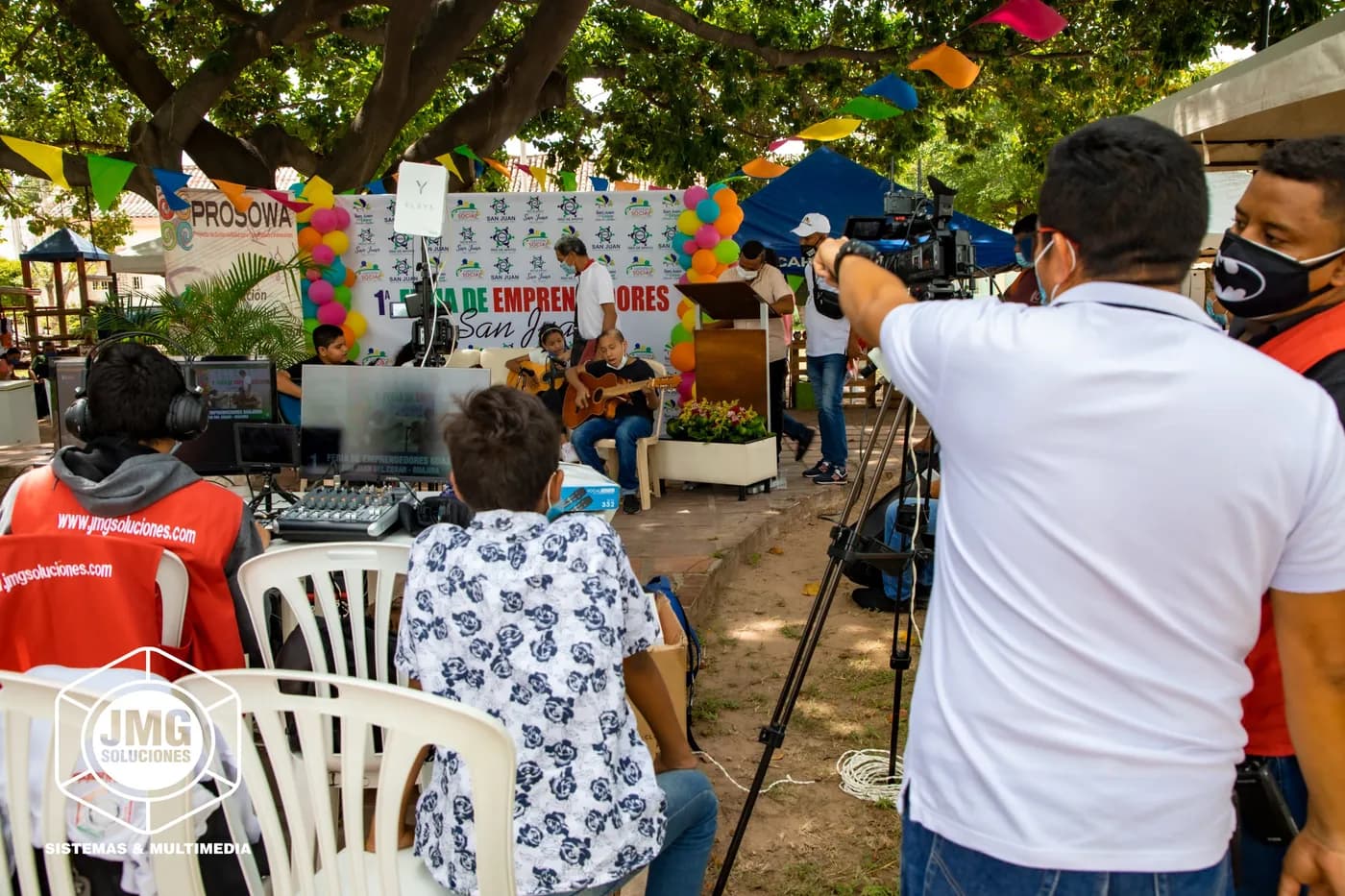 Equipo de JMG transmite una feria de emprendedores al aire libre, con cámara sobre trípode y consola bajo los árboles.