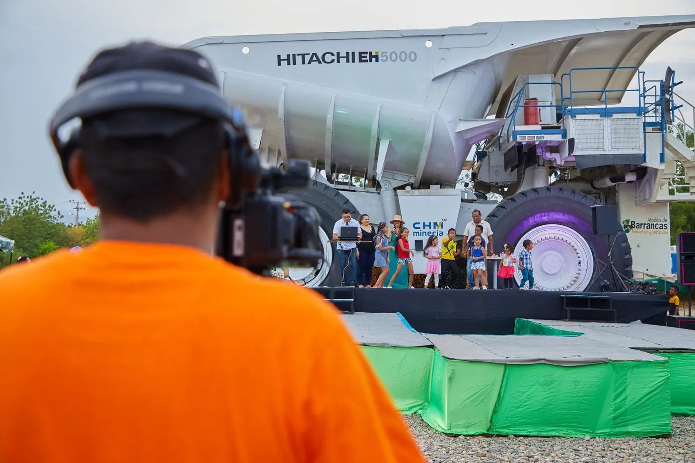 Camarógrafo de JMG con camiseta naranja graba un evento en la tarima frente al camión minero Hitachi EH5000 de Barrancas.