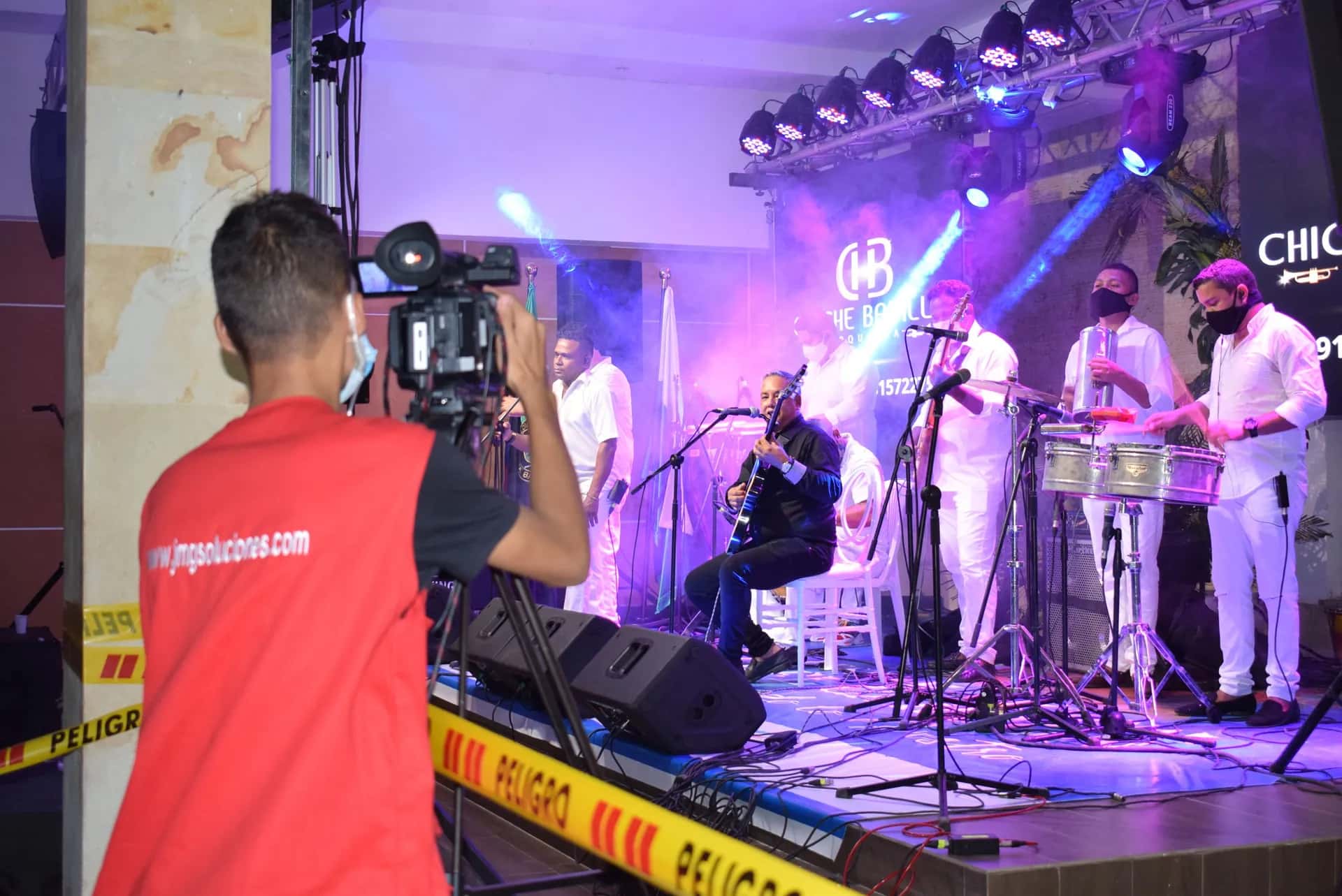 Camarógrafo de JMG con chaleco rojo graba un concierto de música en vivo, con el escenario iluminado en tonos morados.