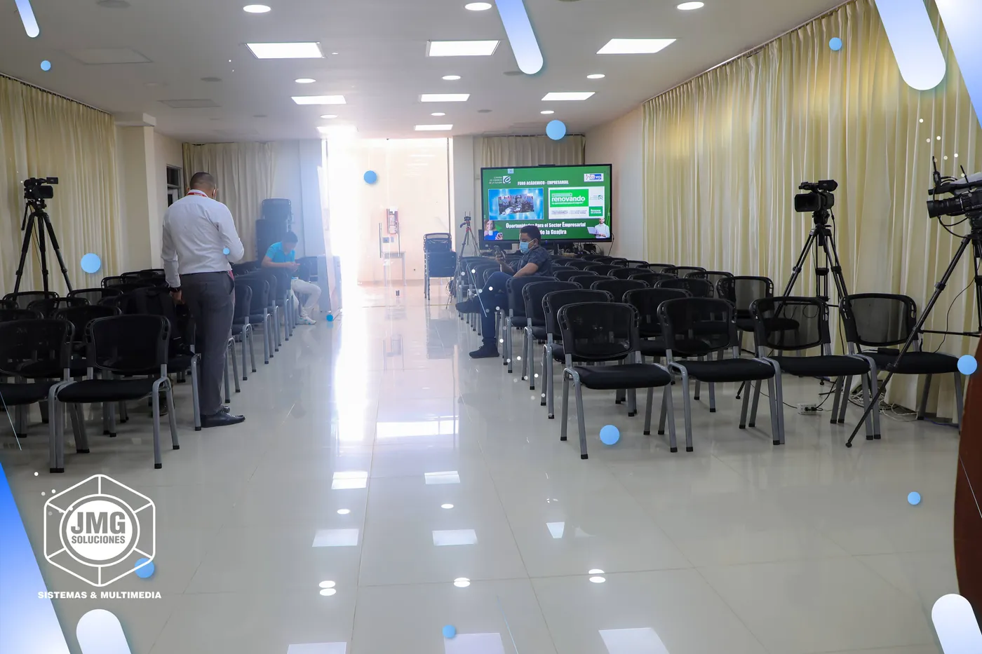 Auditorio preparado por JMG para una transmisión, con cámaras sobre trípodes entre las sillas y una pantalla al fondo.