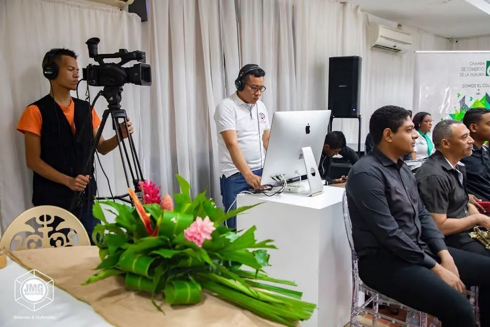 Camarógrafo y operador de JMG transmiten un acto institucional con invitados sentados frente a la mesa de control.