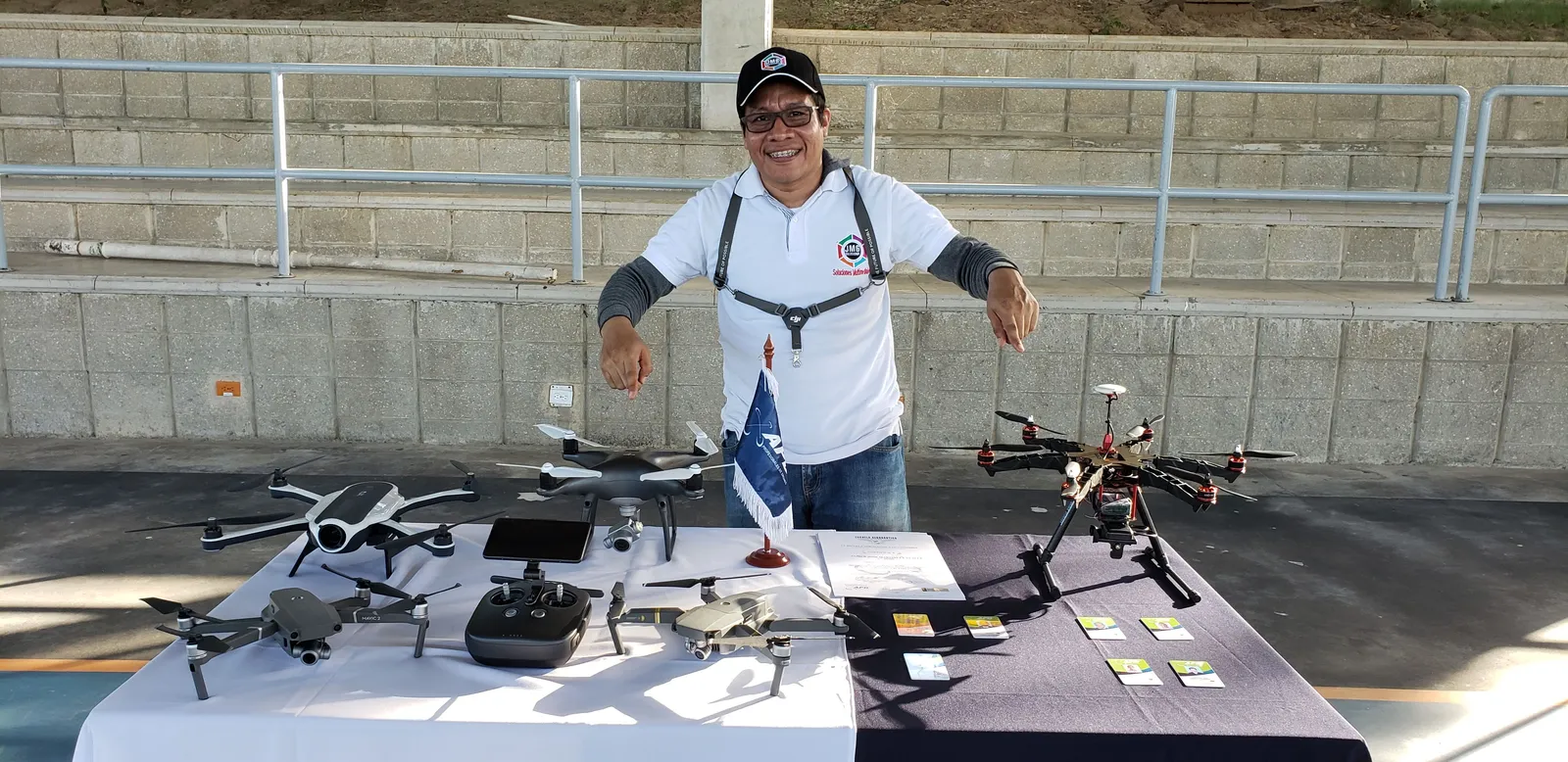 Jaider Mengual sonríe y señala con ambos brazos los drones de una mesa de exhibición.